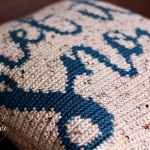 K&ouml;nnte beinhalten: Nahaufnahme eines geh&auml;kelten Kissens mit einem geometrischen Muster in Beige- und T&uuml;rkist&ouml;nen. Das Kissen hat eine strukturierte Oberfl&auml;che und tr&auml;gt ein kleines Logo mit dem Text "TWO little birds patterns".