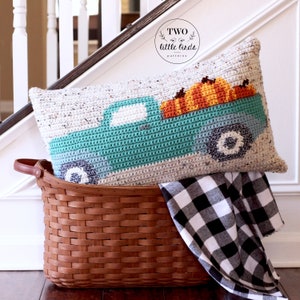 May include: A crocheted cushion featuring a teal blue pickup truck carrying a load of orange pumpkins. The cushion is in a brown wicker basket with a black and white checked fabric.