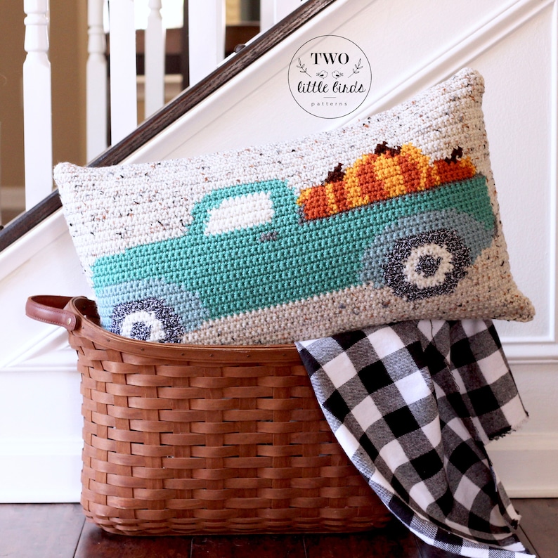 May include: A crocheted cushion featuring a teal blue pickup truck carrying a load of orange pumpkins. The cushion is in a brown wicker basket with a black and white checked fabric.