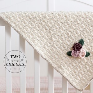 May include: A cream-colored crocheted baby blanket with a floral applique in shades of pink and brown. The blanket is draped over a white crib.