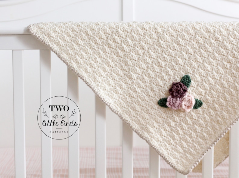 May include: A cream-colored crocheted baby blanket with a floral applique in shades of pink and brown. The blanket is draped over a white crib.