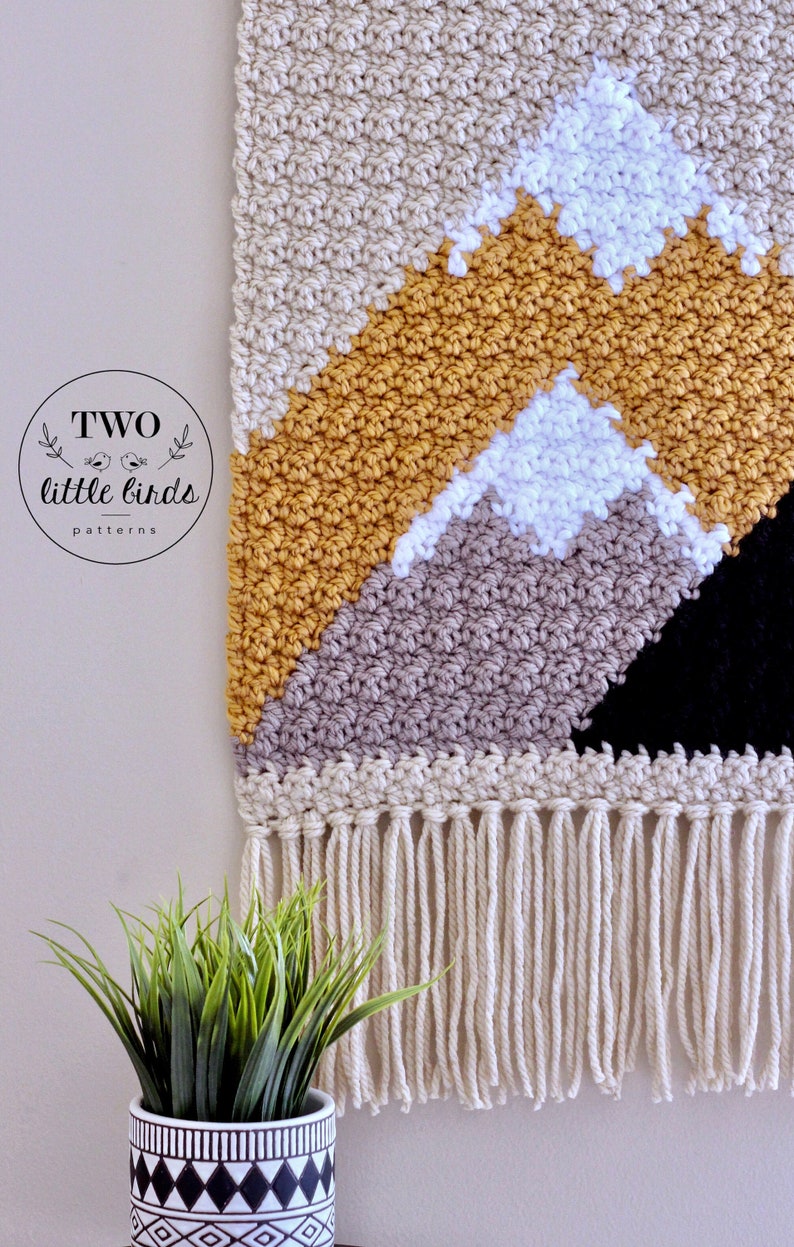 May include: A beige, gray, white, yellow, and black crocheted wall hanging with a fringe border. The wall hanging features a geometric mountain design. The text "TWO little birds patterns" is in a circle at the top left corner of the image.
