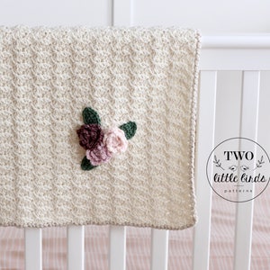 May include: A cream-colored crocheted baby blanket with a floral applique in shades of pink and green. The blanket is hanging on a white crib with a pink and white striped sheet. The text "TWO little birds patterns" is visible in a circle on the crib.
