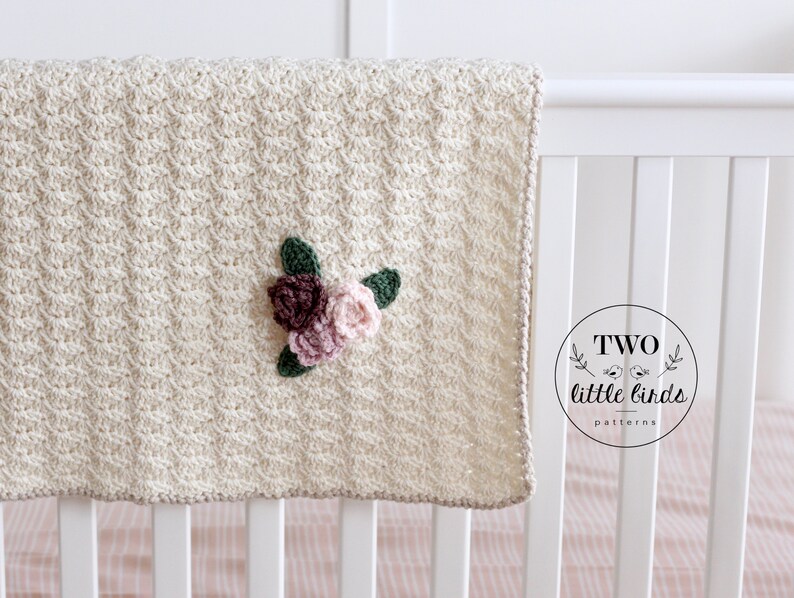 May include: A cream-colored crocheted baby blanket with a floral applique in shades of pink and green. The blanket is hanging on a white crib with a pink and white striped sheet. The text "TWO little birds patterns" is visible in a circle on the crib.