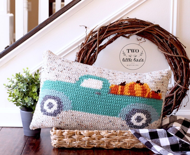May include: A crocheted cushion featuring a teal truck carrying pumpkins. The cushion is placed on a wicker basket and a black and white checked fabric. The text "TWO little birds patterns" is on a brown wreath behind the cushion.