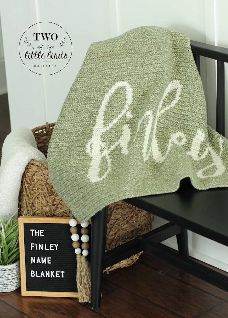 May include: A sage green knitted blanket with the name "Finley" in white script is draped over a black chair. A woven basket and a sign that reads "THE FINLEY NAME BLANKET" are in the background. The blanket is a cosy home decor item.