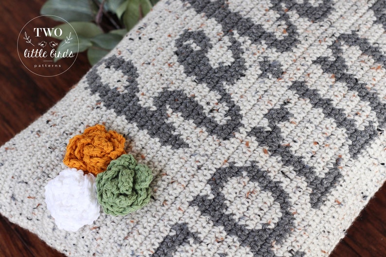 May include: A gray and white crocheted pillow with a geometric pattern. Three crocheted flowers are attached to the pillow: one white, one green, and one orange.