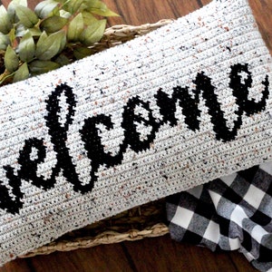 May include: A white crocheted pillow with black yarn spelling out the word "Welcome". The pillow is sitting on a woven basket with a black and white checkered fabric in the background.