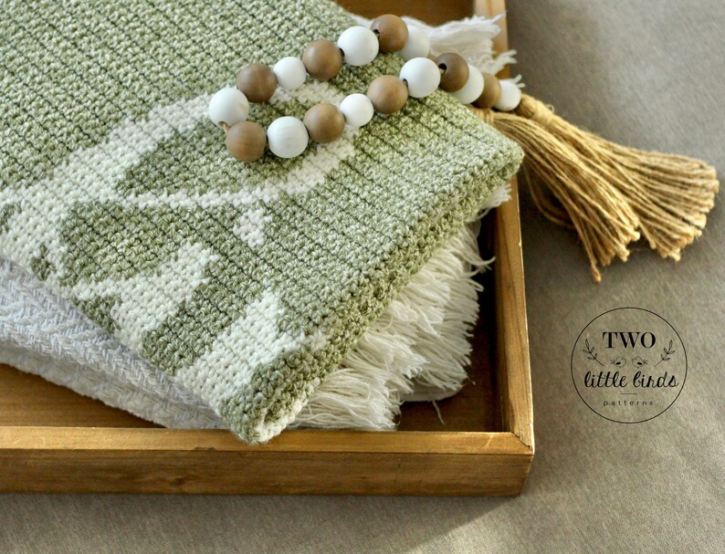 May include: Two crocheted blankets in a wooden tray. The top blanket is light green with a white geometric pattern and fringe. A beaded garland with white and brown beads and a tassel are on the tray. The text "TWO little birds patterns" is in the bottom right corner.