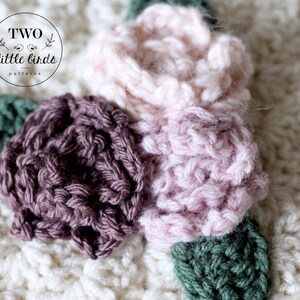 May include: Close-up of a crocheted flower applique with three flowers in shades of pink and brown, and green leaves. The flowers are made with a textured stitch.