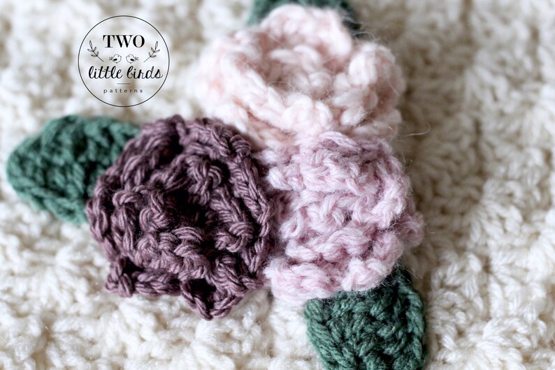 May include: Close-up of a crocheted flower applique with three flowers in shades of pink and brown, and green leaves. The flowers are made with a textured stitch.