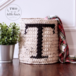 Op de afbeelding: Een gehaakte opbergmand met een grote zwarte letter "T" aan de voorkant. De mand is beige en heeft een gespikkelde textuur. Hij staat op een houten vloer met een geruite deken ernaast.