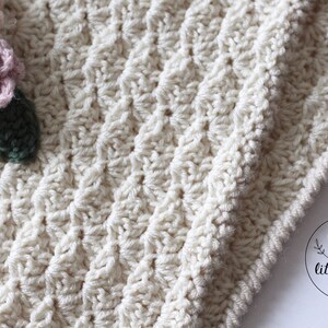 May include: A close-up of a crocheted blanket with a textured stitch pattern. The blanket is off-white and has a crocheted flower embellishment in shades of pink, green, and brown. The image includes the text "TWO little birds patterns".