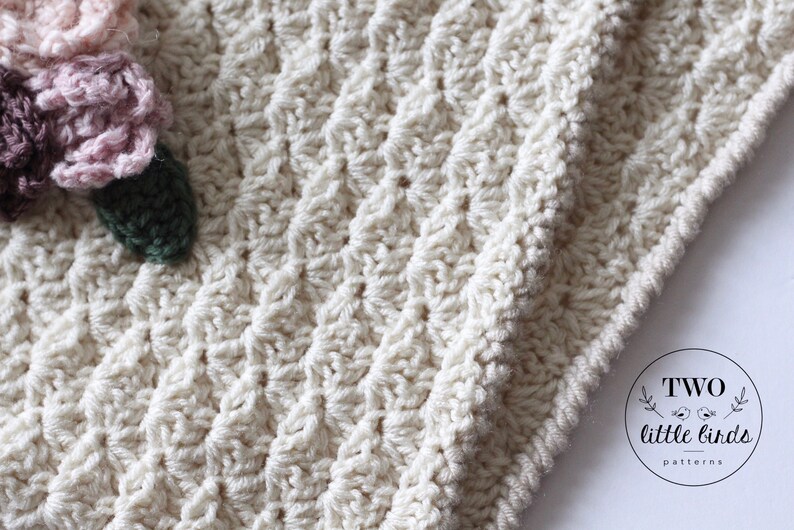 May include: A close-up of a crocheted blanket with a textured stitch pattern. The blanket is off-white and has a crocheted flower embellishment in shades of pink, green, and brown. The image includes the text "TWO little birds patterns".