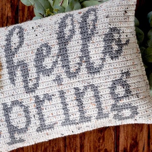 May include: A crocheted pillow with the text "Hello Fall" in gray yarn on a white background. The pillow is decorated with three crocheted flowers in white, green, and orange.