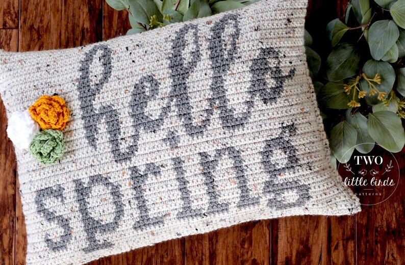 May include: A crocheted pillow with the text "Hello Fall" in gray yarn on a white background. The pillow is decorated with three crocheted flowers in white, green, and orange.