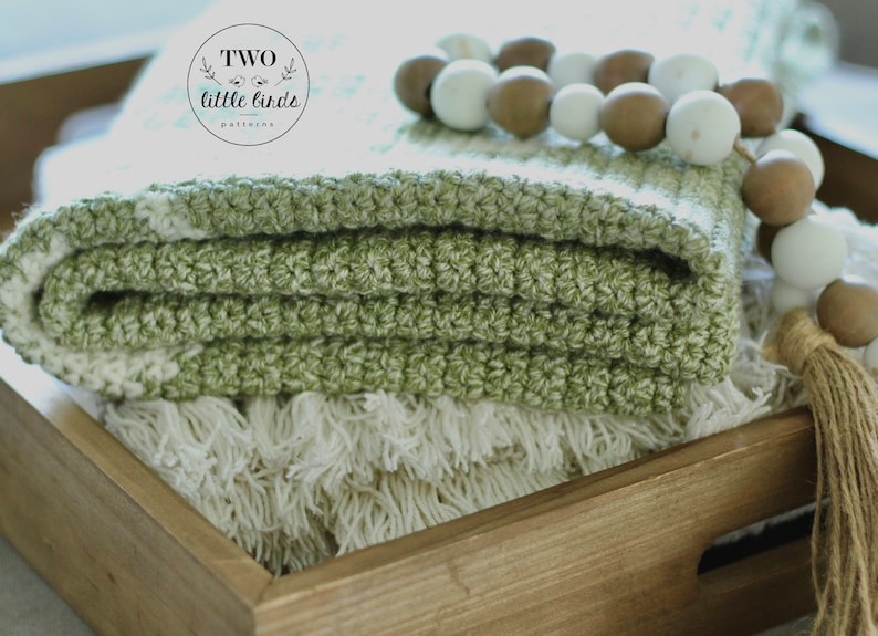 May include: A folded green and white crochet blanket sits in a wooden tray, with a cream-coloured fringed blanket beneath. A beaded garland with white and brown beads and a jute tassel decorates the scene. The text "TWO little birds patterns" is visible.