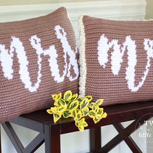Op de afbeelding: Twee bruine gehaakte kussens met witte letters. De kussens zeggen "Mr." en "Mrs.". De kussens liggen op een donkerbruine houten tafel met een klein boeket gele bloemen ervoor.