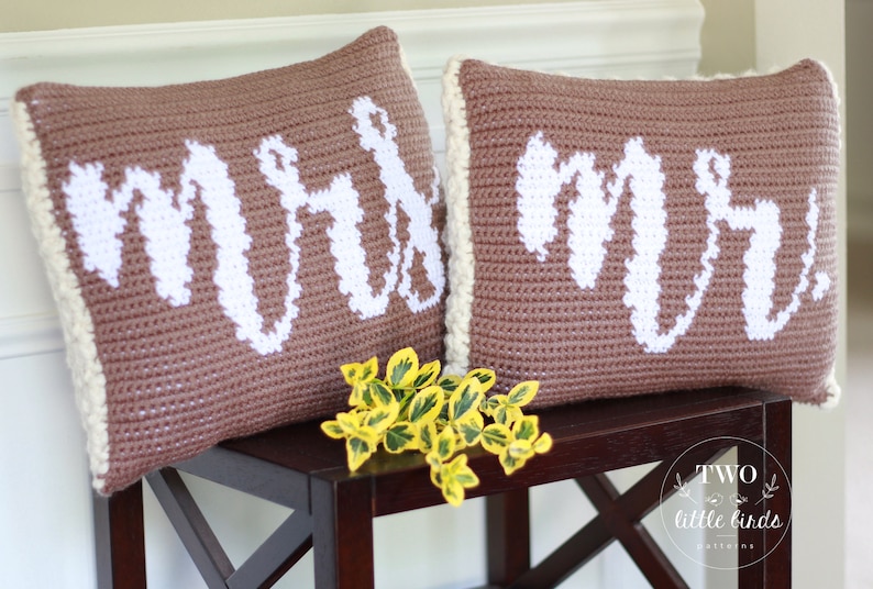 Op de afbeelding: Twee bruine gehaakte kussens met witte letters. De kussens zeggen "Mr." en "Mrs.". De kussens liggen op een donkerbruine houten tafel met een klein boeket gele bloemen ervoor.