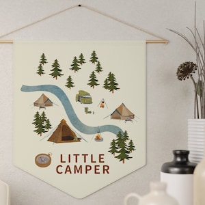 May include: A cream-colored fabric wall hanging with a camping scene illustration. The design includes tents, trees, a river, and a compass. The words "LITTLE CAMPER" are printed in brown. The hanging is suspended from a wooden rod.