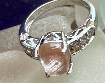 May include: A silver ring featuring a central oval-shaped peach-colored sunstone cabochon. The band is crafted with intricate openwork metal detailing on the shoulders and is accented with small clear stones set along the sides of the ring shank.