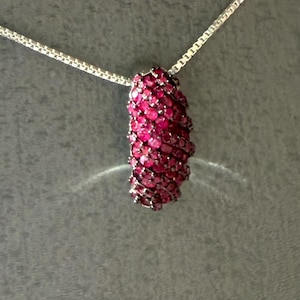 May include: A silver necklace with a pendant. The pendant is a cylindrical shape, covered in small, vibrant red gemstones. The chain is a delicate, silver box chain. The background is a solid, dark gray.