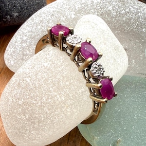 Könnte beinhalten: Ein goldfarbener Ring mit ovalen, tiefroten Edelsteinen und kleinen, runden, klaren Edelsteinen. Der Ring liegt auf einem weißen Stein, mit anderen Steinen und einer Holzoberfläche im Hintergrund.