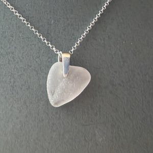 May include: A silver necklace featuring a heart-shaped, frosted glass pendant. The pendant hangs from a delicate silver chain. The necklace is displayed on a grey mannequin.