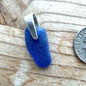 May include: A deep blue sea glass pendant with a silver loop. A UK ten pence coin is next to the pendant for scale. The sea glass has a smooth, rounded form. The background is a light-coloured wooden surface.