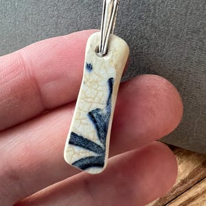 May include: A rectangular pendant necklace with a silver chain. The pendant is off-white with a blue floral design. The pendant is approximately 2.5 cm long.