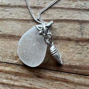 May include: A silver necklace with a frosted, teardrop-shaped glass pendant. A small, silver, cone-shaped charm hangs beside the glass. The delicate chain supports the pendant, which is attached with a silver bail.