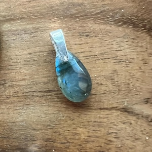 May include: A teardrop-shaped pendant with a silver bail, showcasing a blue-green iridescent gemstone. The pendant is set against a wooden surface, highlighting the natural colors and textures of the stone.