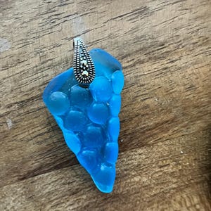 May include: A blue pendant with a silver bail. The pendant has a unique shape and a textured surface. The bail is decorated with small, round silver accents. The pendant is displayed on a wooden surface.