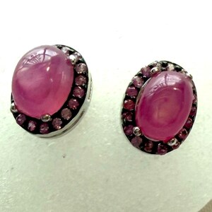 Puede incluir: Un par de pendientes de botón con una piedra central rosa de corte cabujón ovalado, rodeada por un halo de pequeñas piedras rosas engastadas en metal oscuro. El engaste oscuro contrasta con el vibrante color rosa de las piedras.