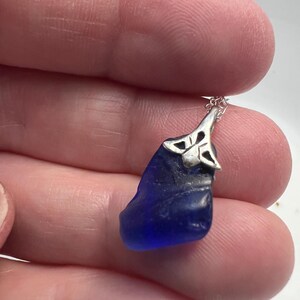 May include: A dark blue sea glass pendant with a silver-coloured Celtic knot design. The pendant hangs from a delicate silver chain. The sea glass has a smooth, rounded shape.