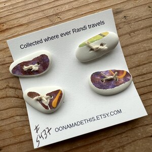 May include: Four unique ceramic buttons with different designs and colors. The buttons are attached to a white card with the text "Collected where ever Randi travels" and "OONAMADETHIS.ETSY.COM".