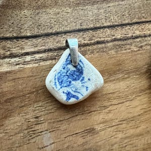 May include: A pendant crafted from a piece of sea pottery. The pendant has a light base with a blue floral design and a silver-coloured bail. The pendant is an irregular shape, perfect for a necklace.