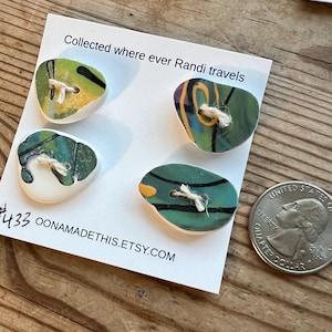 May include: Four ceramic buttons with green, black, and yellow abstract designs. Each button has a hole in the center and a white string threaded through it. The buttons are displayed on a white card with the text "Collected where ever Randi travels" and "#5433 OONAMADETHIS.ETSY.COM".