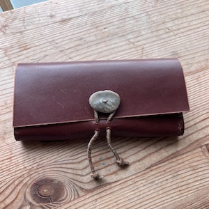 May include: A burgundy leather wallet with a stone clasp and a knotted closure. The wallet is rectangular and appears to be folded over. The background is a wooden surface.