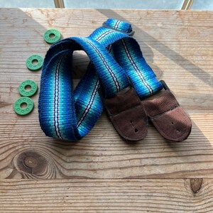May include: A blue and turquoise guitar strap with brown leather ends and a striped pattern. Three green guitar picks with star designs are also visible on a wooden surface.