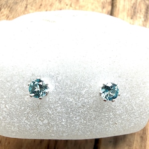 May include: A pair of stud earrings featuring round, faceted blue-green gemstones set in silver-toned metal prongs. The earrings are displayed against a smooth, white, rounded surface, highlighting the clarity and color of the stones.