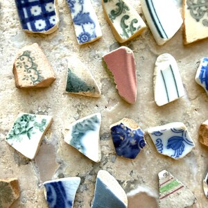 May include: A collection of antique ceramic shards with various patterns and colors. The fragments feature blue and white floral designs, green swirls, and pink and white accents. The pieces are arranged on a light-colored surface.