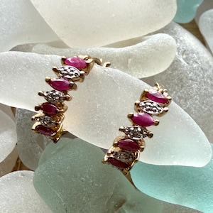 May include: A pair of gold hoop earrings with alternating ruby and diamond accents. The earrings are set against a backdrop of sea glass in shades of white, blue, and green.