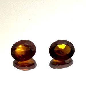 Op de afbeelding: Twee ovaalvormige, gefacetteerde edelstenen met een diepe amber- en oranje tint. Elke steen heeft een gepolijst oppervlak dat licht reflecteert, waardoor een helder, gloeiend centrum ontstaat. De edelstenen zijn naast elkaar geplaatst.