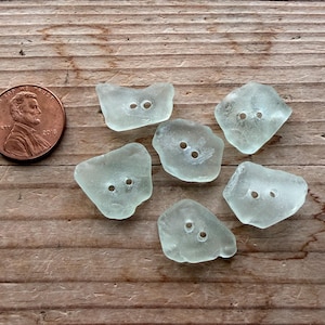 May include: Six sea glass buttons in various shapes and sizes, each with two holes for sewing. The buttons are a pale, translucent green color. A penny is next to the buttons for size comparison.