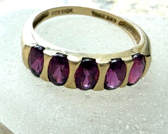 May include: A gold-toned ring featuring five oval-cut deep purple gemstones set in a row. The interior of the band is engraved with the text 925, SCB, and THAILAND. The metal band has a smooth, polished finish and a curved, solid construction.