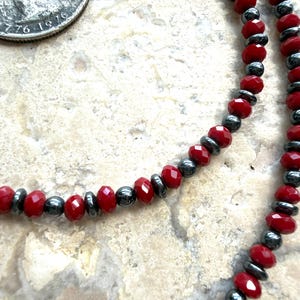 May include: A beaded necklace featuring alternating faceted bright red crystal beads and smooth metallic dark gray hematite-style spacer beads. A silver-colored United States quarter coin is visible in the upper left corner for scale.