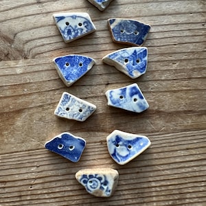 May include: A collection of ten irregular, flat ceramic buttons featuring blue and white patterns. Each button has two small holes for attachment. The pieces exhibit varied geometric shapes with smooth, worn edges, showcasing distinct floral and abstract designs.