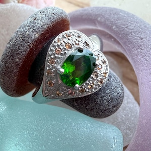 May include: A silver ring with a heart-shaped setting. The setting is encrusted with small, light brown stones and surrounds a large, oval-cut, vibrant green gemstone. The ring is displayed on a bed of colorful, smooth stones.