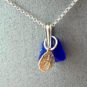 May include: A silver chain necklace with a pendant. The pendant has a piece of blue sea glass and a small gold-coloured leaf charm. The pendant hangs from a silver bail. The background is grey.